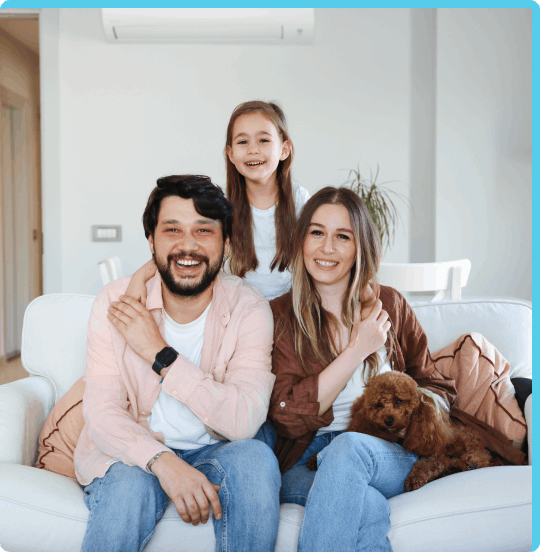 Family smiling and sitting on a white couch with a small dog with an AC unit visible on the wall behind them