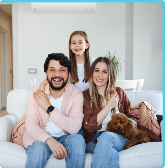 Family smiling and sitting on a white couch with a small dog with an AC unit visible on the wall behind them